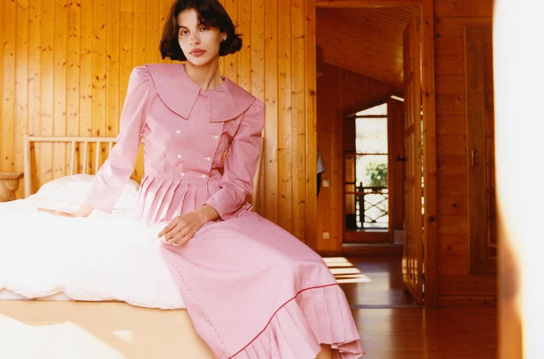 A woman with short brunette hair sits on a bed in a wood-paneled room wearing a pink and white patterned dress, indoors with natural light.