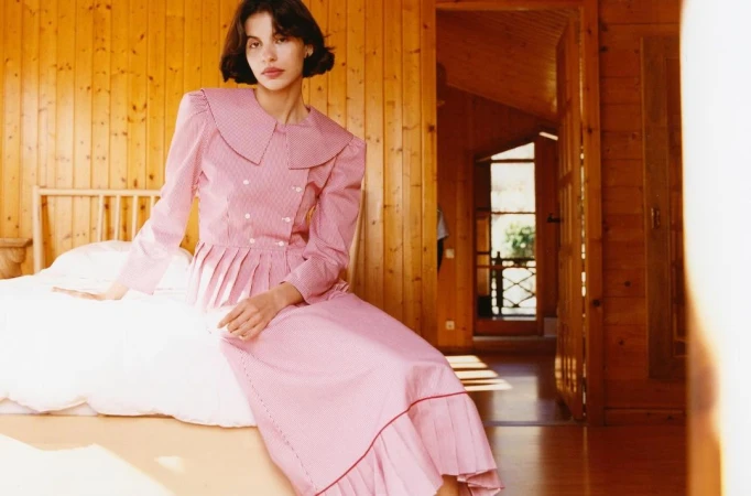 A woman with short brunette hair sits on a bed in a wood-paneled room wearing a pink and white patterned dress, indoors with natural light.