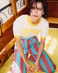 A woman with short dark hair wearing a white ringer t-shirt and striped trousers kneeling indoors by a wooden window.