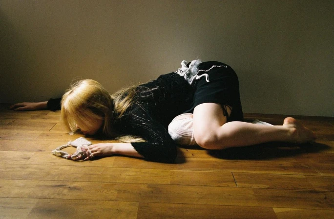 Blonde woman lying curled on a wooden floor wearing black clothing by Alba Yruela