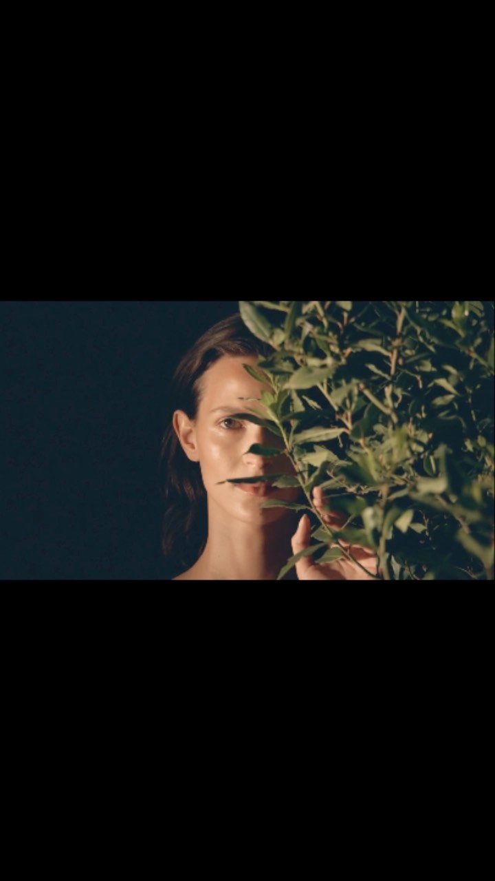 A young woman is partly hidden behind leaves in a moody, close-up shot, illuminated by natural light against a dark background.