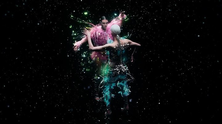 Two models in avant-garde fashion pose against a black backdrop with dispersed particles in an artistic, high-fashion presentation.