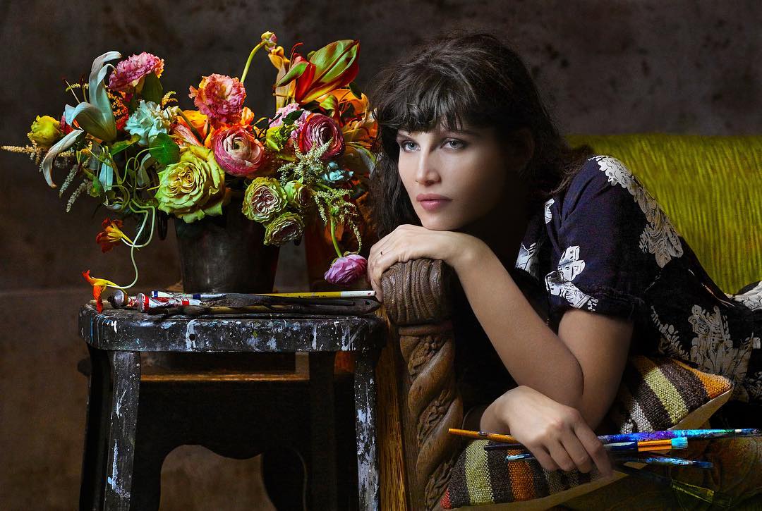 Adult woman lounges beside colorful flower arrangement with paintbrushes and art supplies on a stool in a dimly lit indoor setting.
