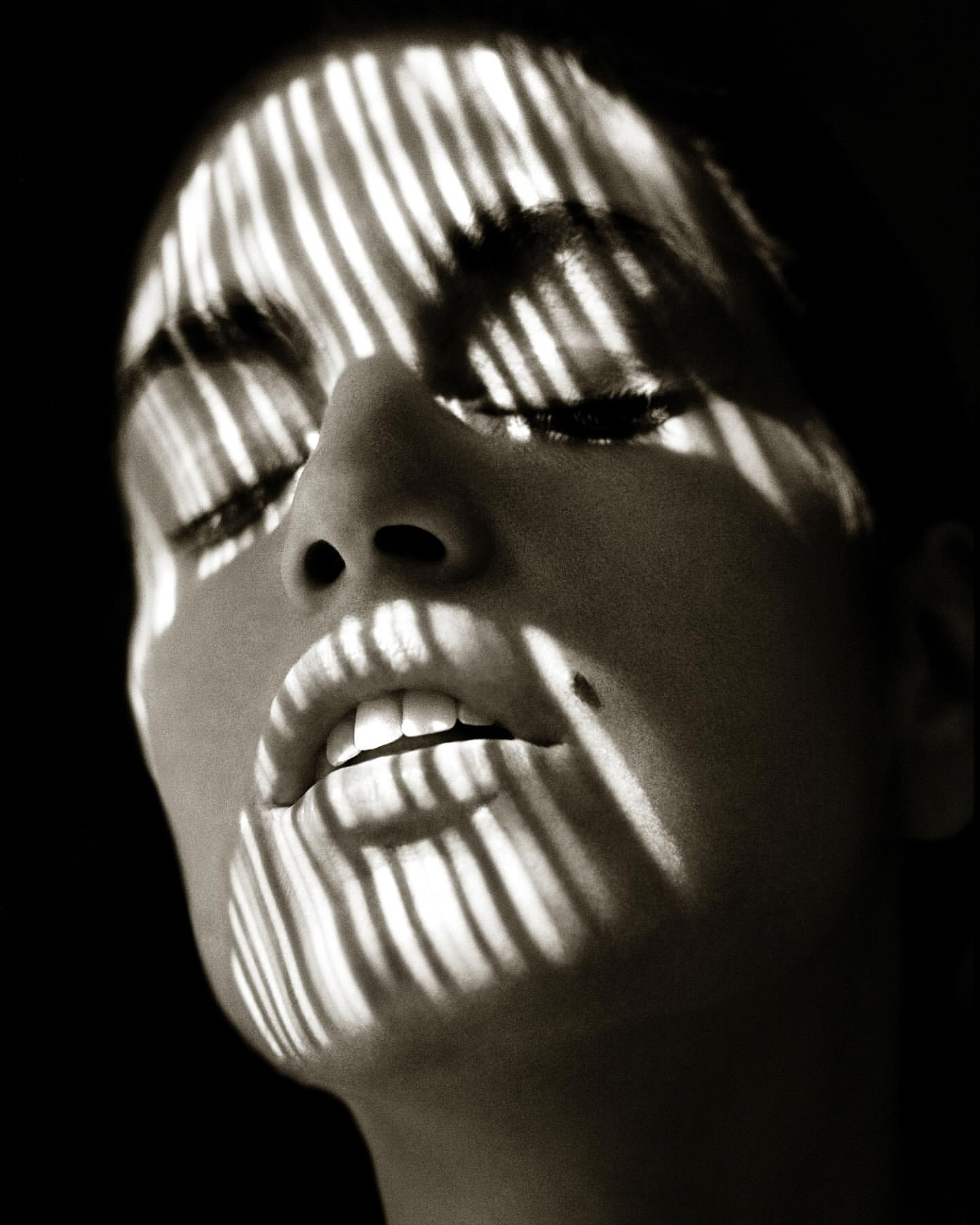 Close-up portrait of a woman's face in black and white with striped lighting across her forehead and eyes in a studio setting.