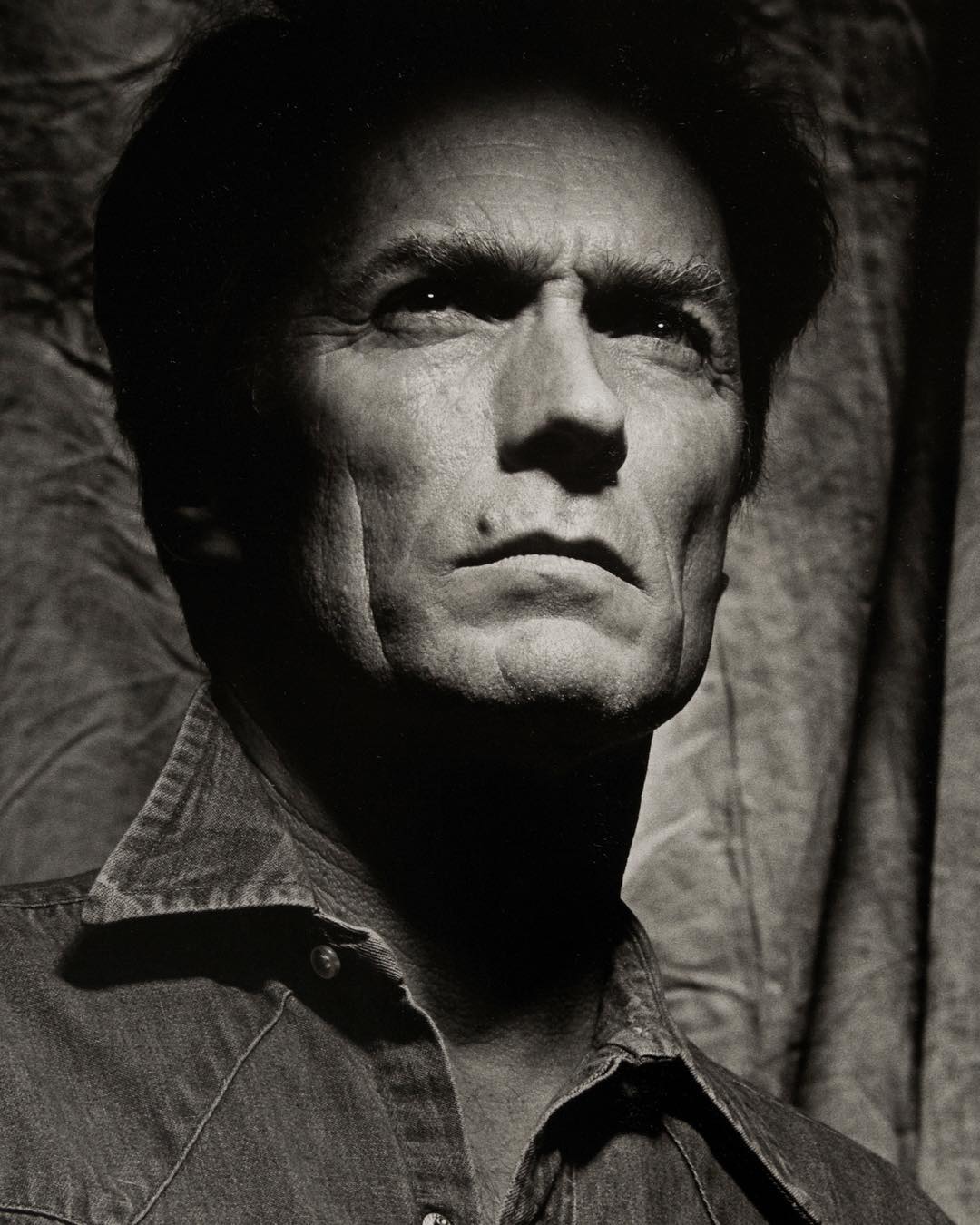 Black and white portrait of adult male with short dark hair wearing a denim shirt, photographed indoors against a draped background.