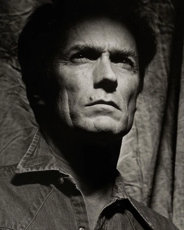 Black and white portrait of adult male with short dark hair wearing a denim shirt, photographed indoors against a draped background.