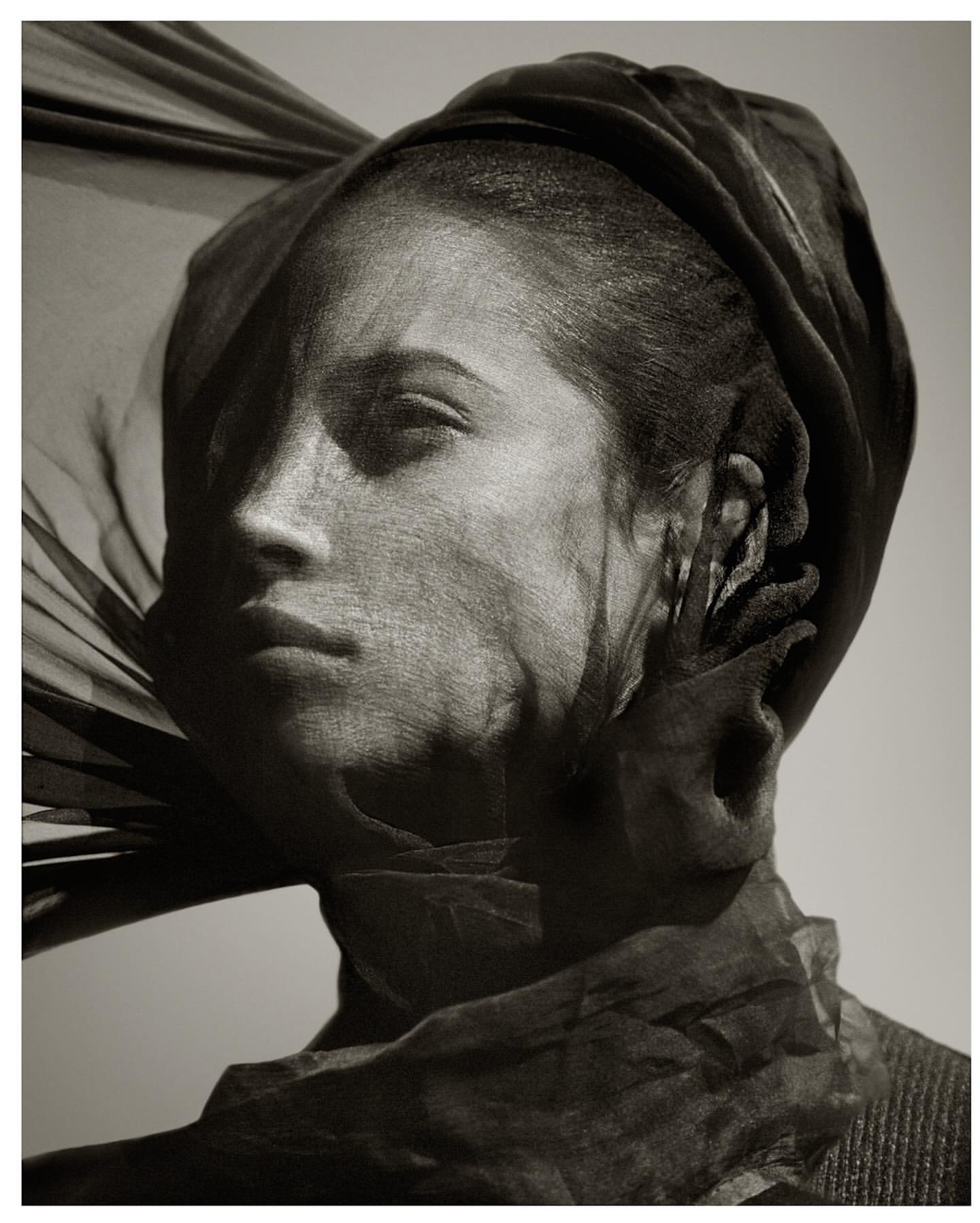 Portrait of an adult female with black cloth draped over her head and face in a studio with a neutral, desaturated background.