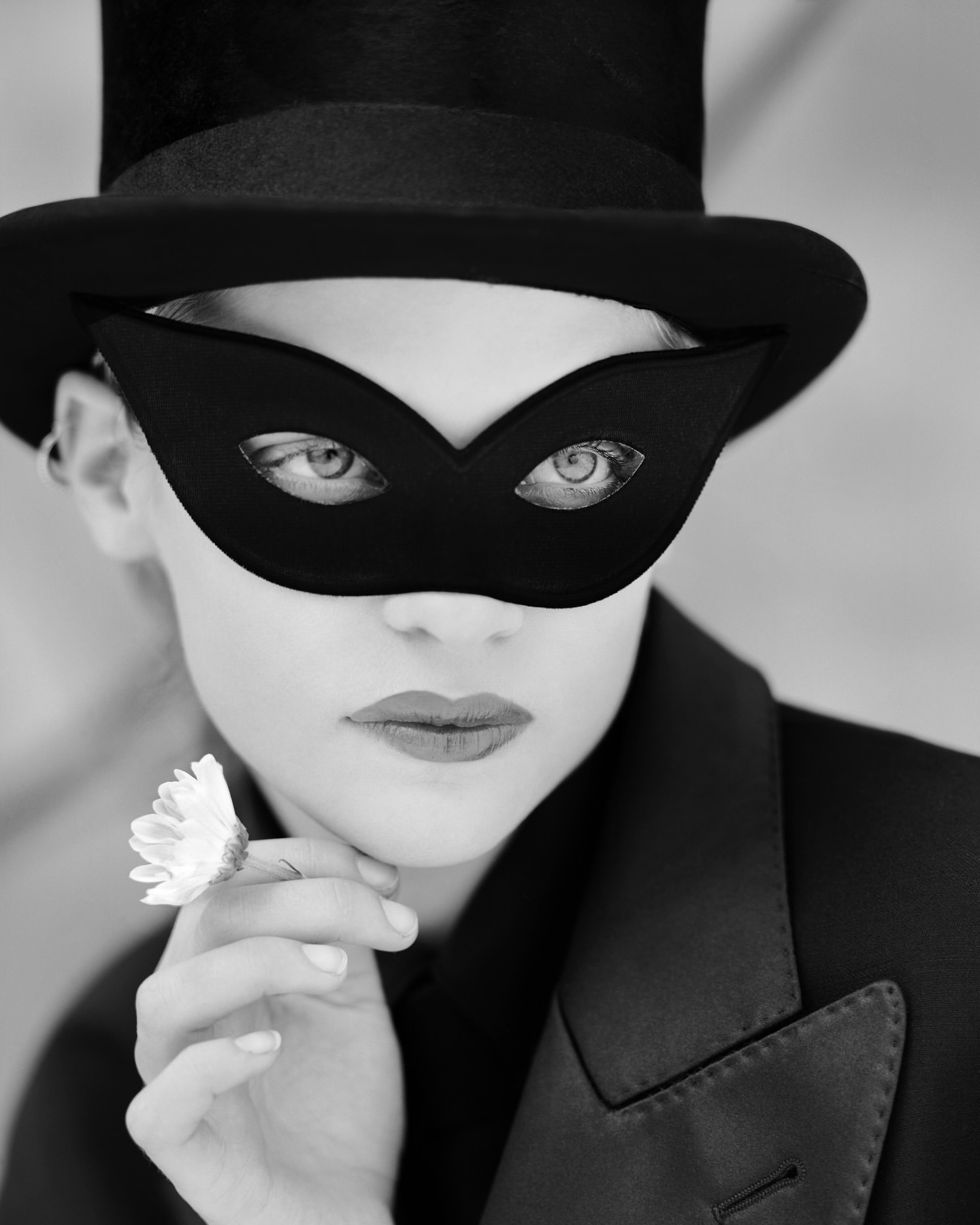 Close-up of a woman wearing a black top hat and mask, holding a white daisy. Black and white portrait in an indoor environment.
