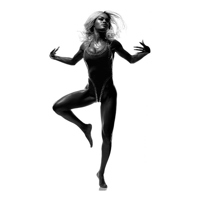 Monochrome portrait of a blond woman in a dark bodysuit, captured in mid-dance against a bright white backdrop in a studio setting.