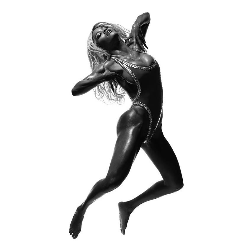 Monochrome image of a woman in a bodysuit leaping with outstretched arms against a stark white background in a studio setting.