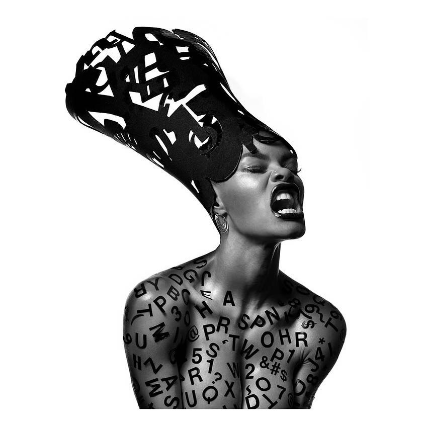 Monochrome close-up studio shot of a Black female with a large textured hat and upper body covered in black typeface.