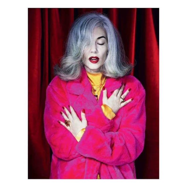 Portrait of an adult woman with gray hair in a pink coat posing in front of red curtains. She is wearing red lipstick and several rings.