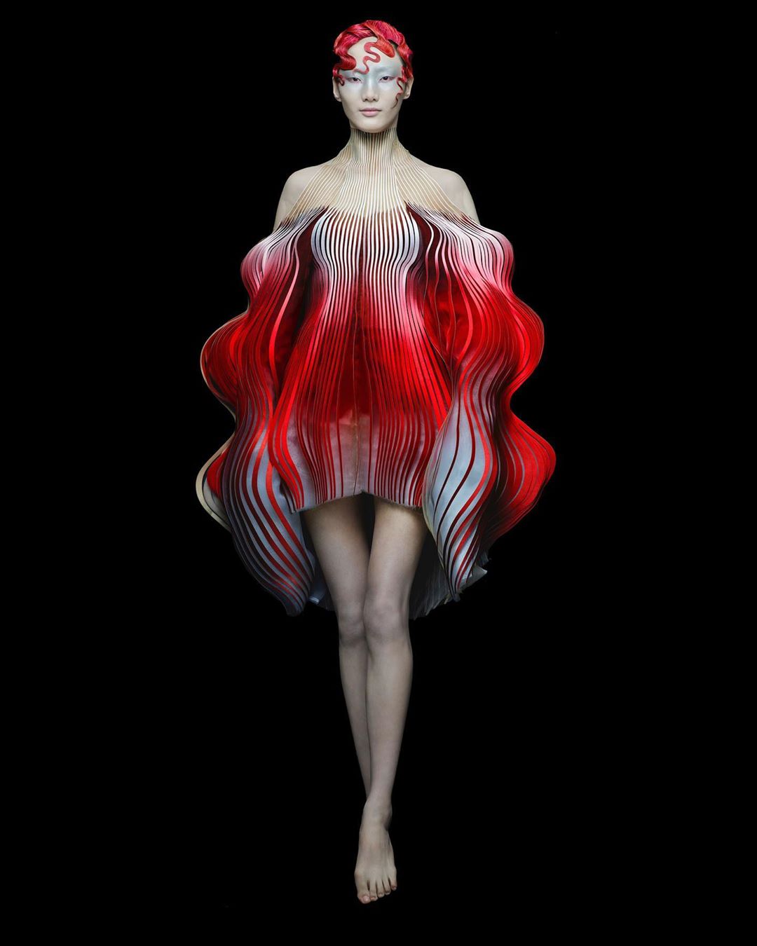 A woman in a futuristic, high-fashion red and white striped dress poses against a stark black backdrop in a studio setting.