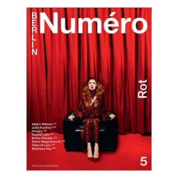 Redheaded woman in shimmering red outfit seated on black armchair against red curtain background for Numéro Berlin magazine