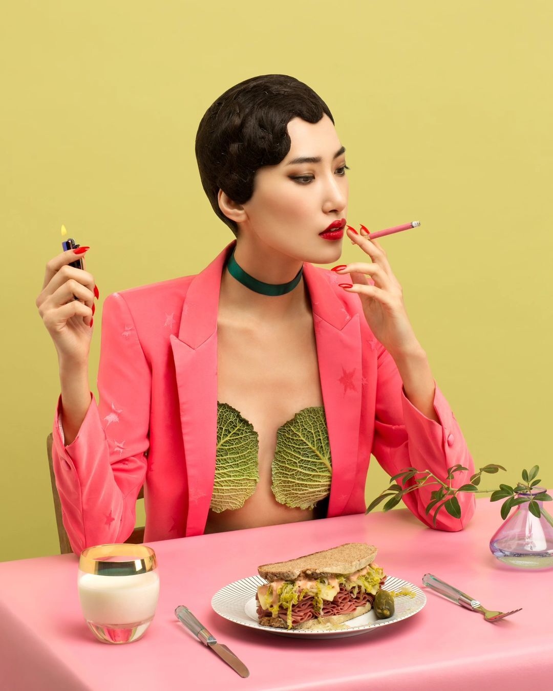 Asian woman in a pink blazer sits at a table with a sandwich, cigarette, and beauty products in a studio with a chartreuse background.