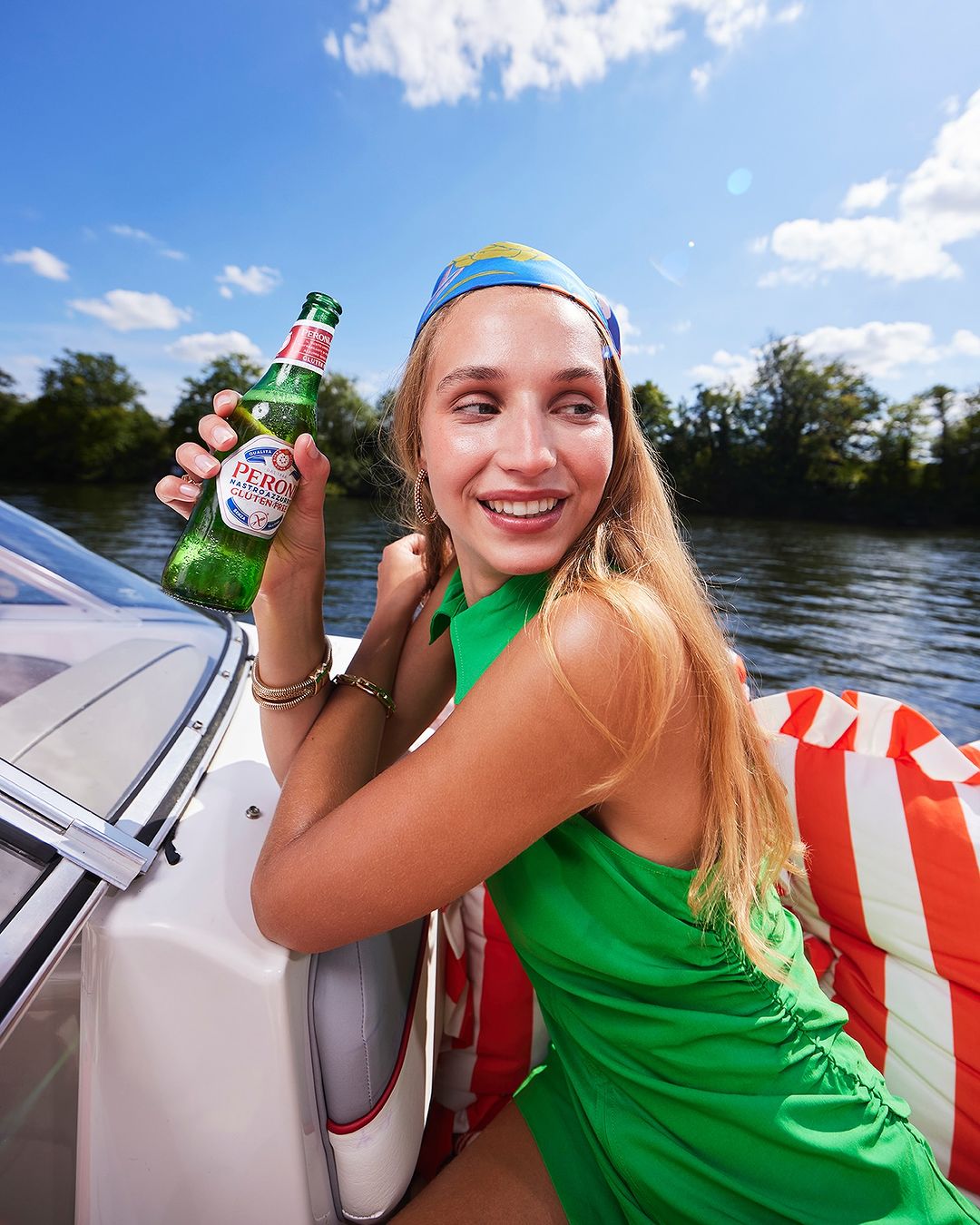 Woman in green dress smiles and holds Peroni beer on a boat under a sunny sky with trees, water, and blue sky.