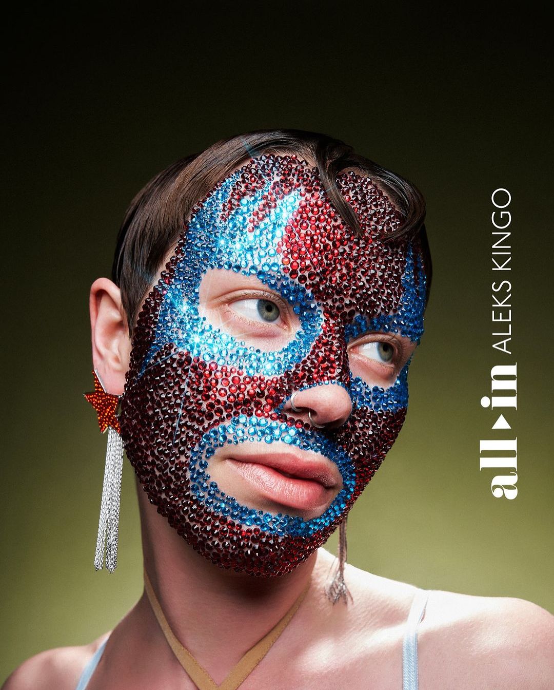 Close-up of a person with a colorful bejeweled mask and star earrings, set against a muted green backdrop, creating a striking beauty image.