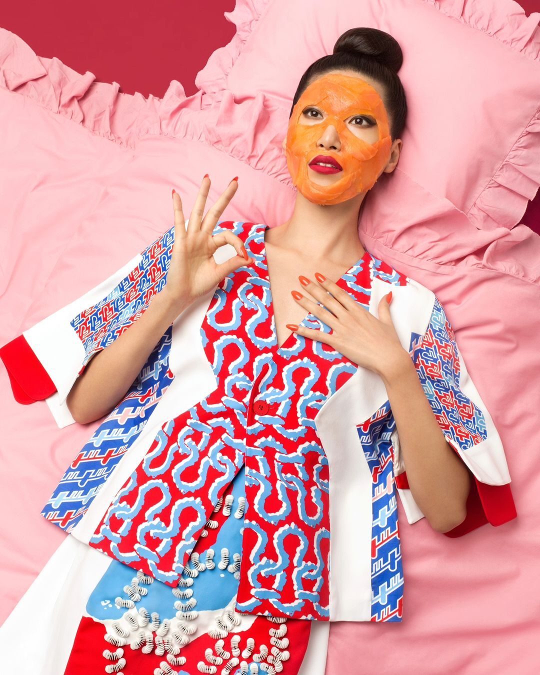 Woman with an orange facial mask lies on pink bedding, wearing a patterned jacket and skirt, posing with an 'OK' hand gesture.