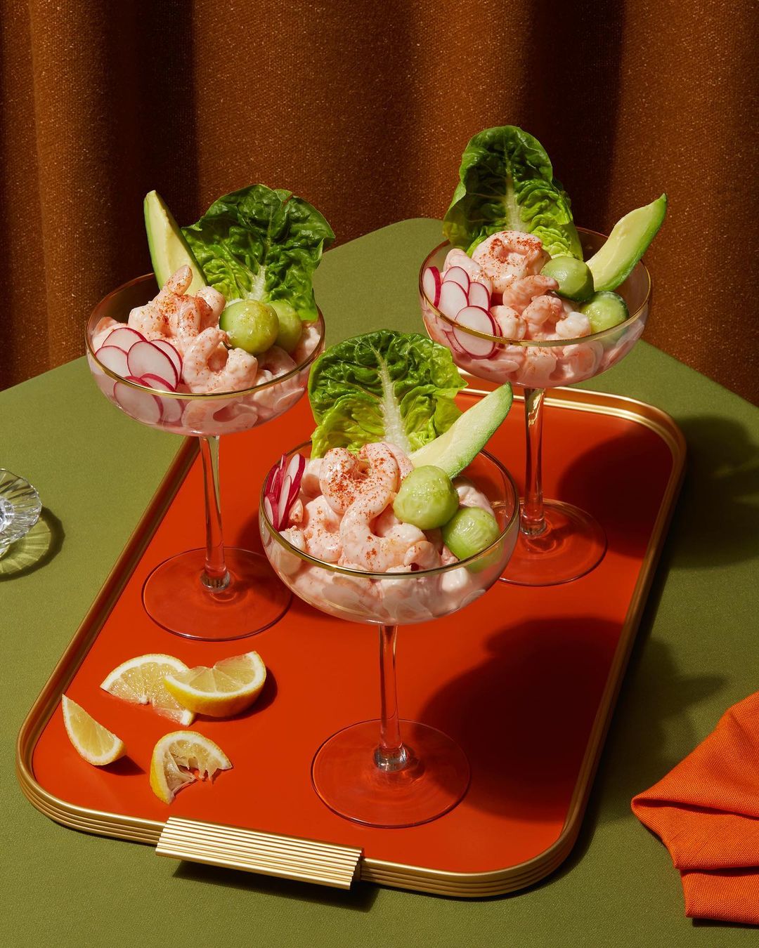 Three shrimp cocktail glasses filled with shrimp, radish, lettuce, and avocado on an orange tray against a brown backdrop.