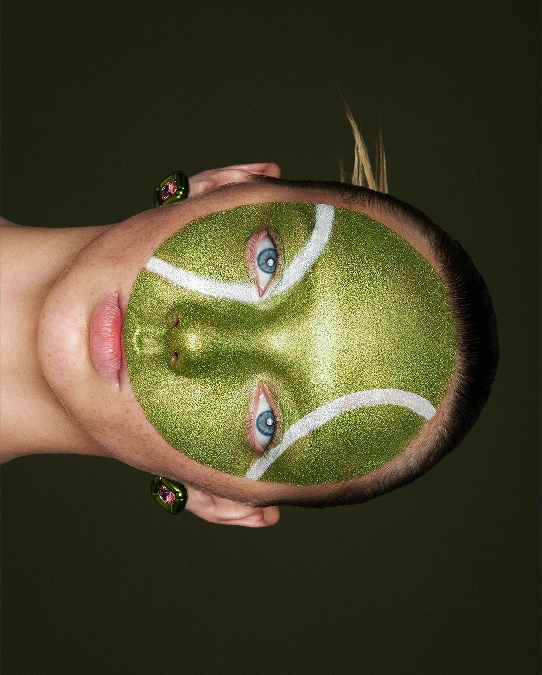 Face of an adult woman with green tennis ball makeup on her face, ring accessory, and neutral background with portrait orientation.
