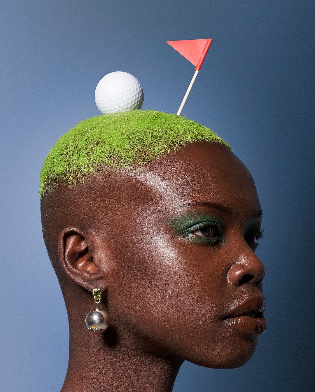 Close-up of Black woman in profile with green hair styled like a golf green topped with a golf ball and flag against a blue gradient background.
