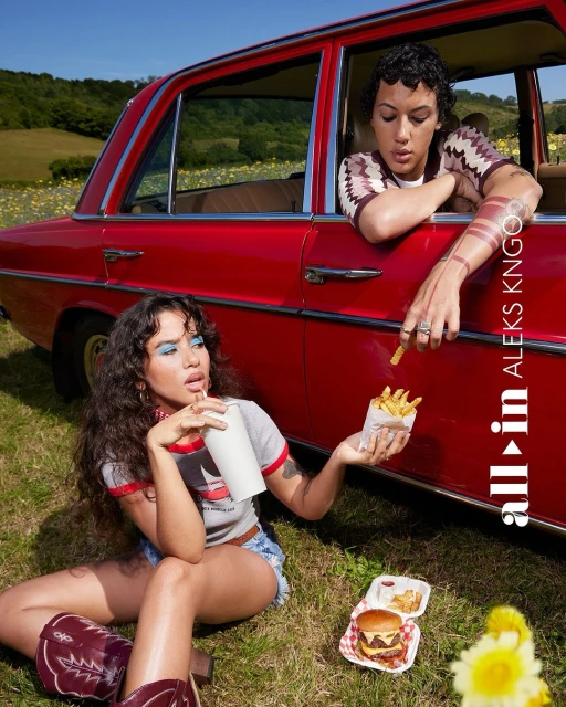 Two women outdoors with a vintage red car, enjoying french fries and a burger on a grassy field under a blue sky with distant hills.