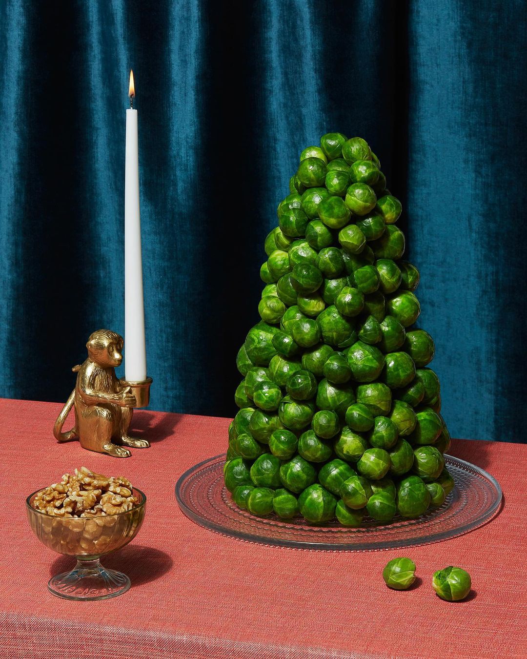 Festive Brussels sprouts Christmas tree sits on a glass plate next to a candle and a bowl of walnuts against a blue curtain backdrop.
