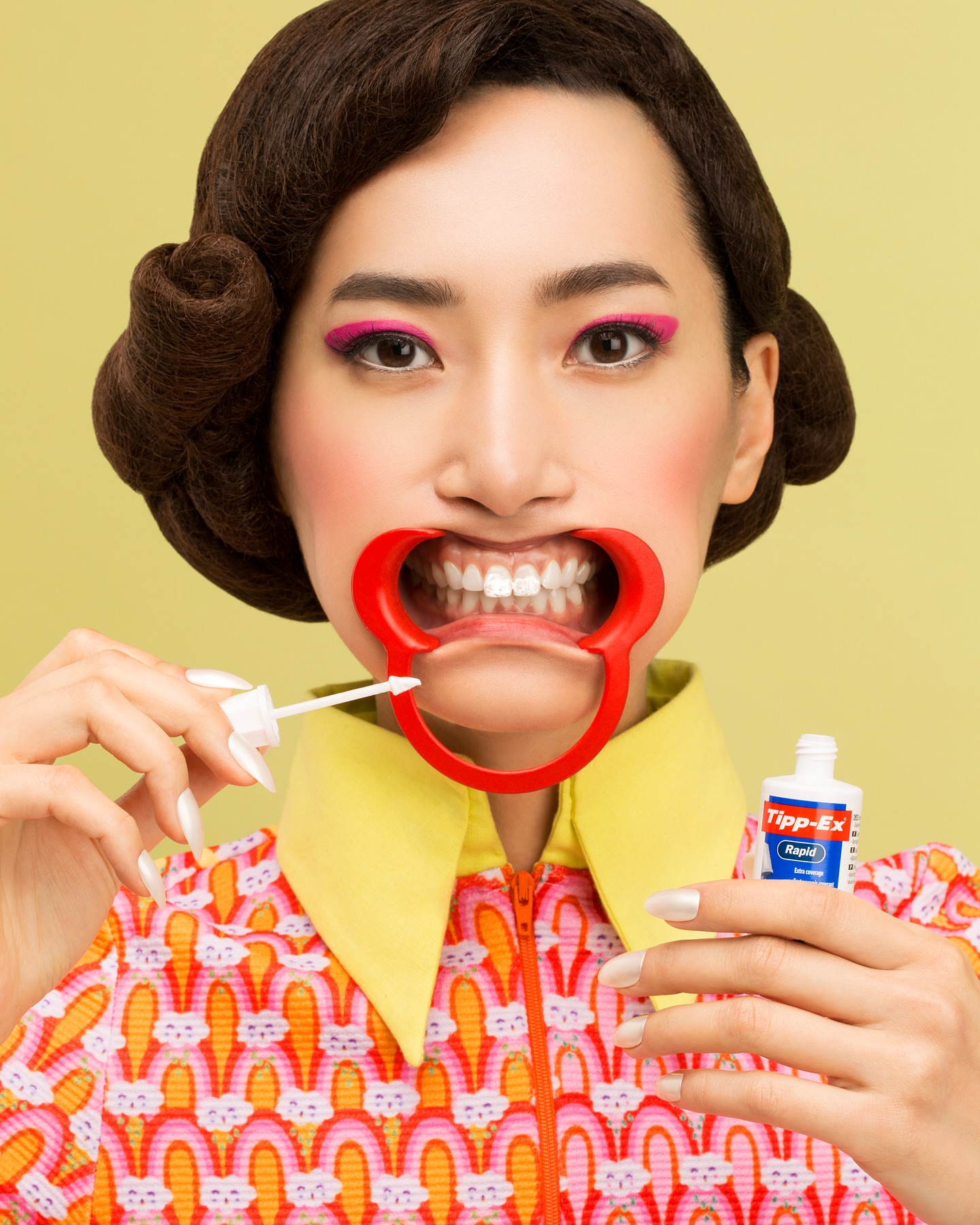 Asian woman with red dental retractor holding white-out. Wearing 60s-style printed dress and hairstyle, standing in front of a yellow backdrop.