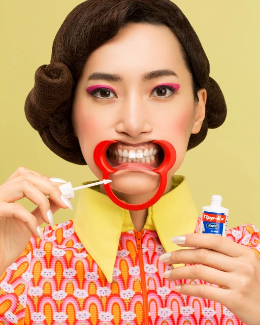 Asian woman with red dental retractor holding white-out. Wearing 60s-style printed dress and hairstyle, standing in front of a yellow backdrop.