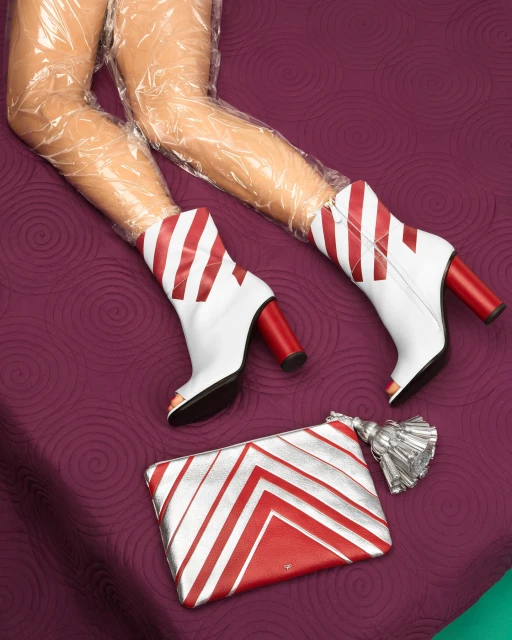 Woman's legs covered in plastic wrap, ankle boots with red heels and stripes, and a red and silver clutch on purple fabric.