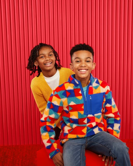 Two smiling children pose in front of a red striped background; one wears a patterned fleece, the other a yellow sweater.