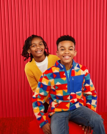 Two smiling children pose in front of a red striped background; one wears a patterned fleece, the other a yellow sweater.