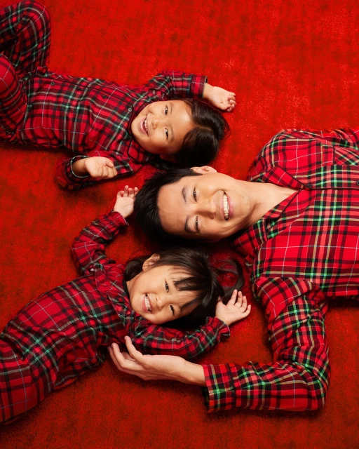 Smiling Asian family, including a man and two young children, wearing matching red and green plaid pajamas laying on a red background.