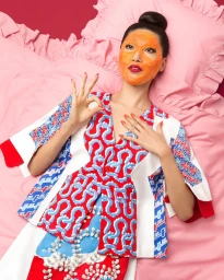 Woman wearing an orange face mask and patterned top reclining on a pink bed by photographer Aleksandra Kingo.