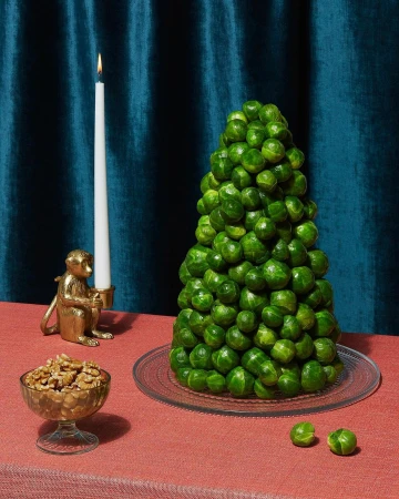 Conical tower of Brussels sprouts on a glass plate next to a lit candle, small golden monkey figurine, and bowl of walnuts on a red tablecloth against deep blue drapes.