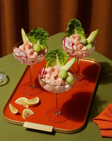 Three shrimp cocktails in stemmed glasses on an orange tray with lemon wedges, avocado slices, and lettuce leaves.