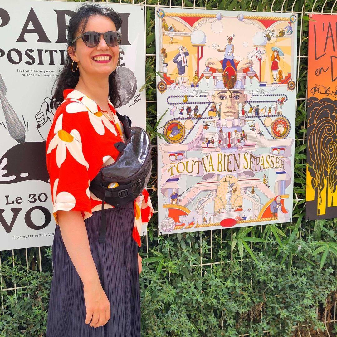 A young woman smiles outdoors wearing a floral shirt and black skirt, posing next to colorful posters with green foliage in the background.