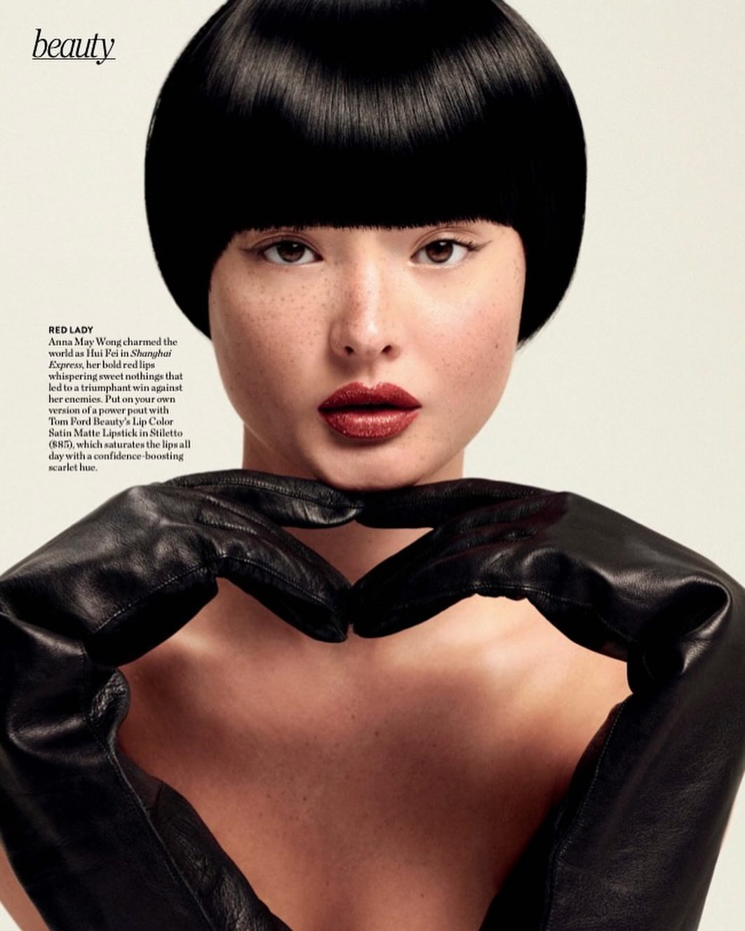 Close-up of an Asian woman with a blunt bob haircut and dark red lipstick, making a heart shape with black leather gloves against a light background.