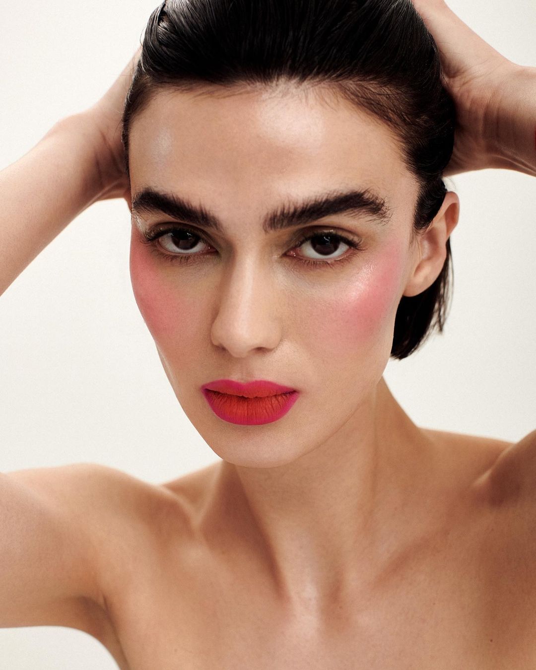 Close-up portrait of a fair-skinned woman with bold makeup, wet-look hair slicked back, bright red lipstick, and pink blush on a white backdrop.