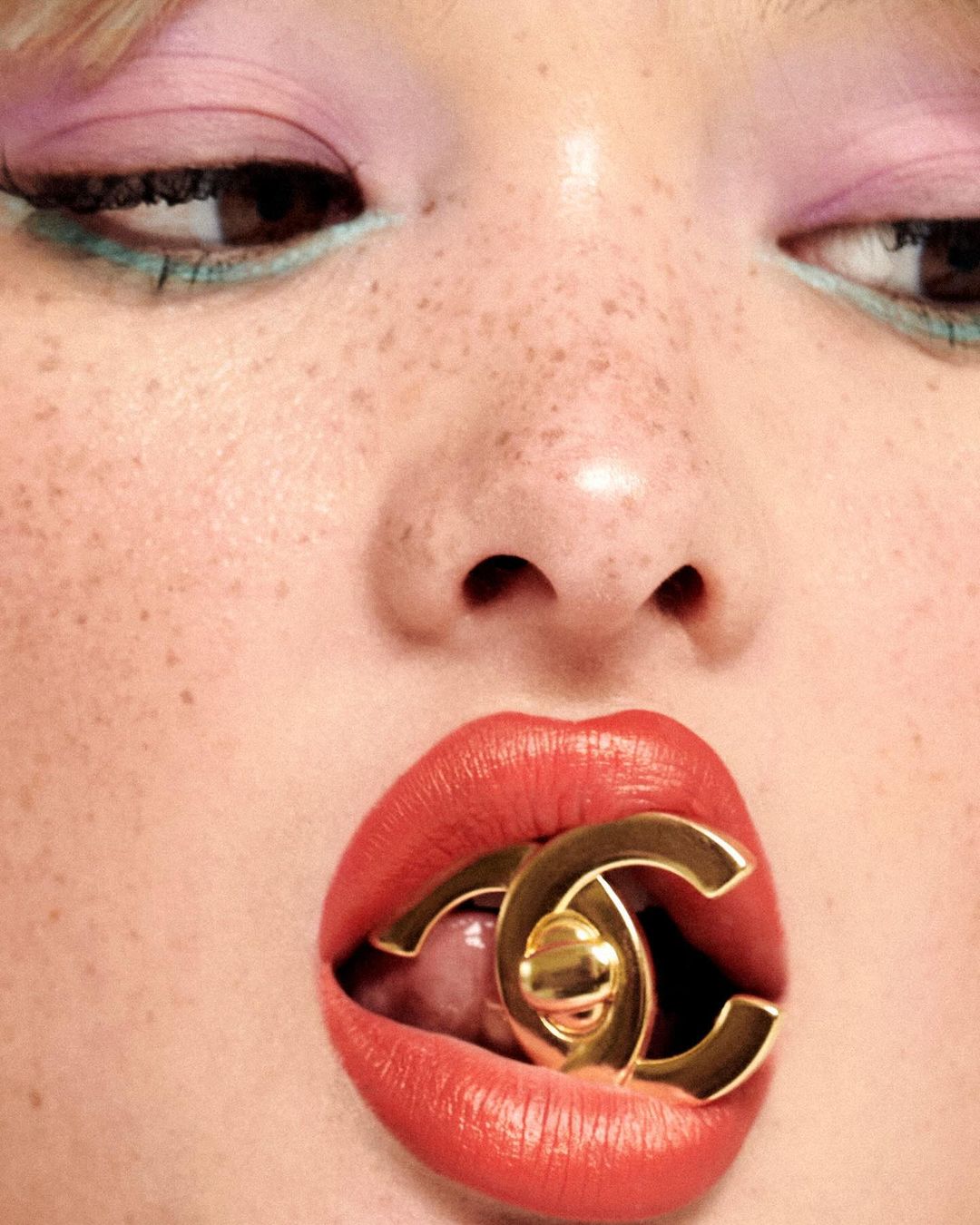 Close-up of a woman with freckled skin, vibrant red lips, and light-colored eye makeup, biting down on a gold 'CC' logo earring.