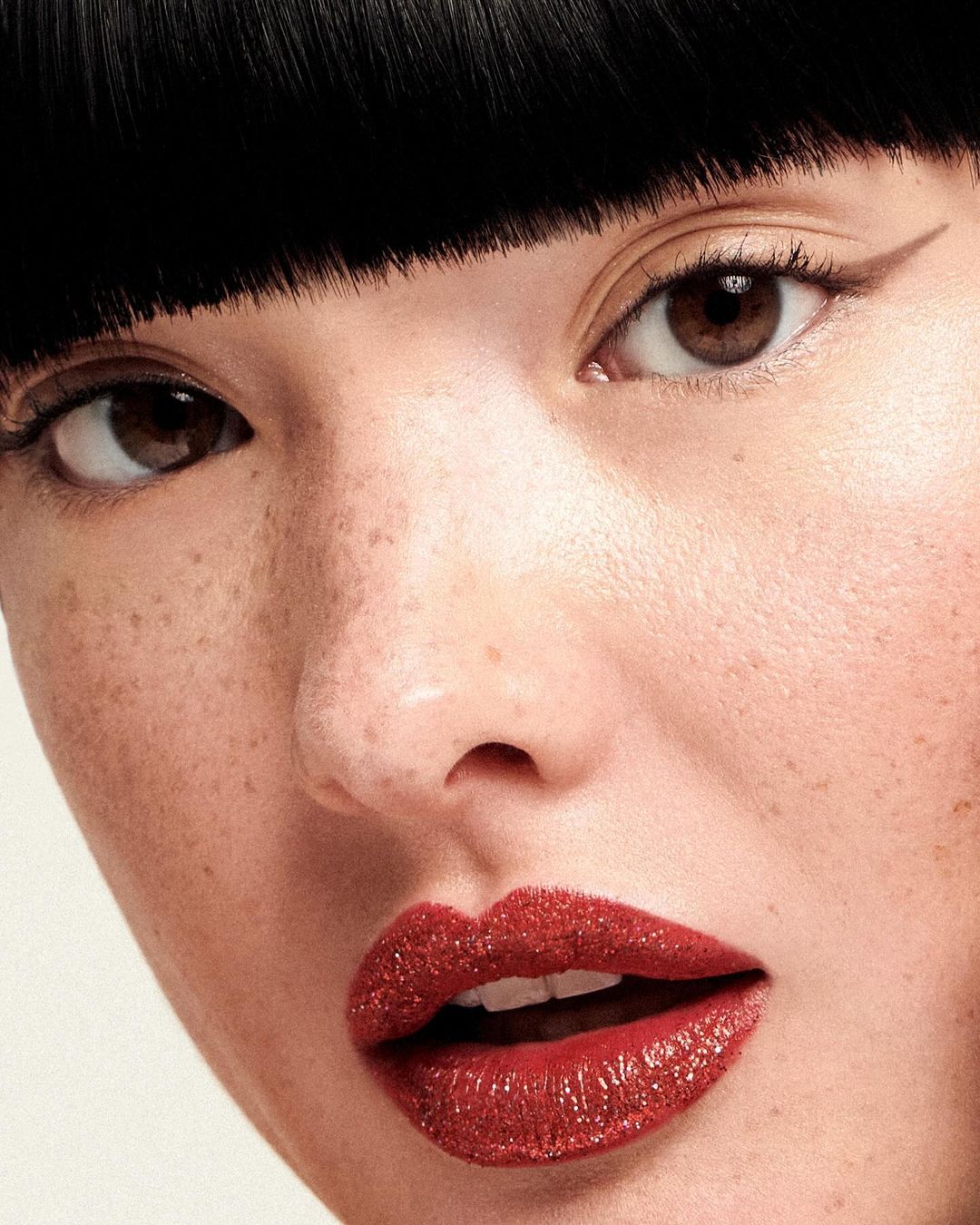 Close-up of a young adult female with glossy red lips and black bangs, showcasing a beauty and fashion aesthetic with a clean background.