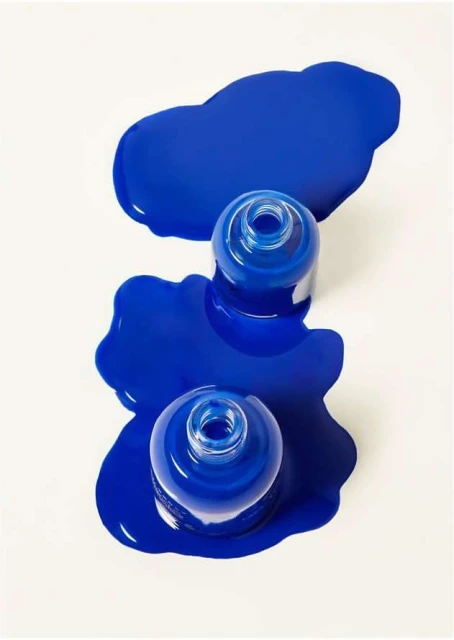 Elevated studio shot showcases three cobalt blue nail polish bottles with liquid spills on a stark white surface. Beauty product.