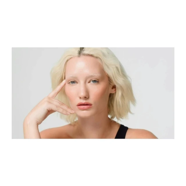 Close-up of a fair-skinned woman with platinum blonde wavy bob, gazing directly at the camera with her hand near her forehead.