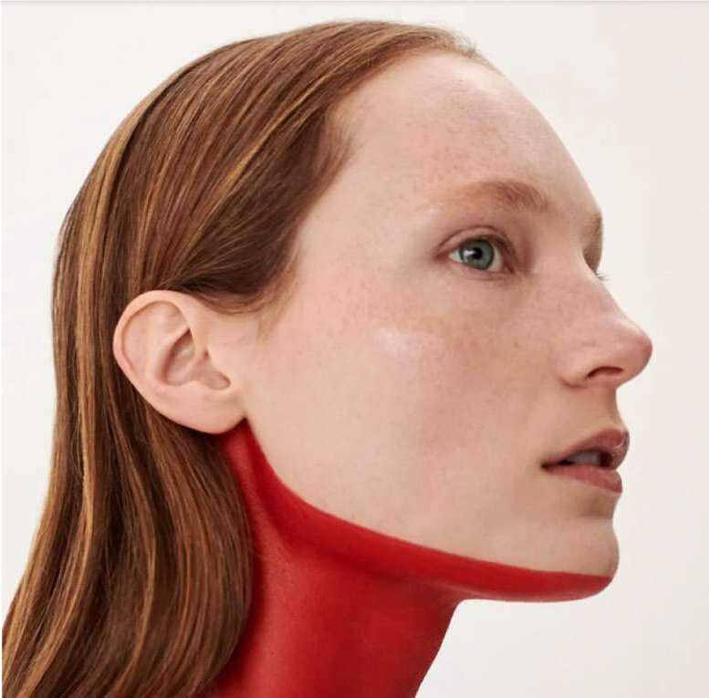 Close-up studio shot of a light-skinned adult female with light brown hair in profile, accented with red paint on the neck and collarbone.