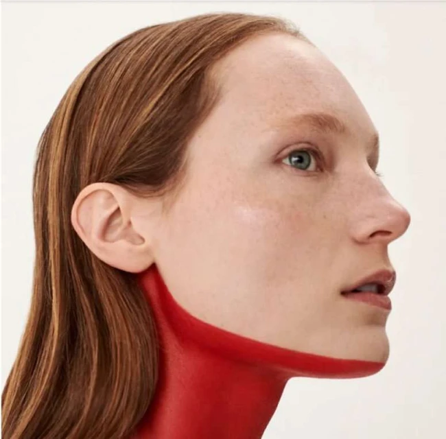 Close-up studio shot of a light-skinned adult female with light brown hair in profile, accented with red paint on the neck and collarbone.