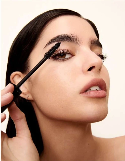 Close-up of a brunette adult woman applying mascara to her eyelashes, with fair skin, soft lighting, and a light background.
