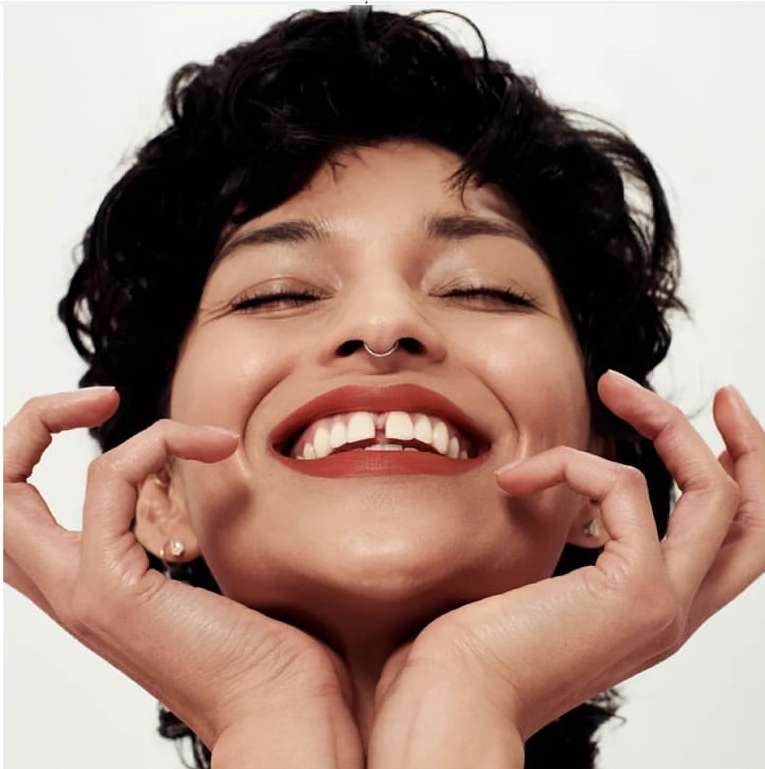 Close-up portrait of a smiling woman with dark curly hair, septum ring, and hands framing her chin, against white seamless background.