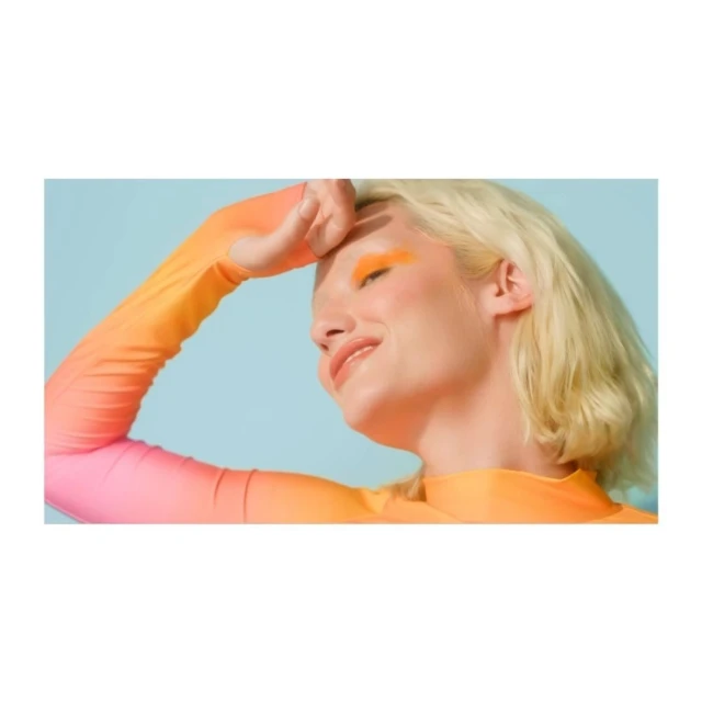 Smiling fair-skinned woman in gradient long-sleeved shirt, shielding eyes with hand, orange eyeshadow, posing against blue backdrop.