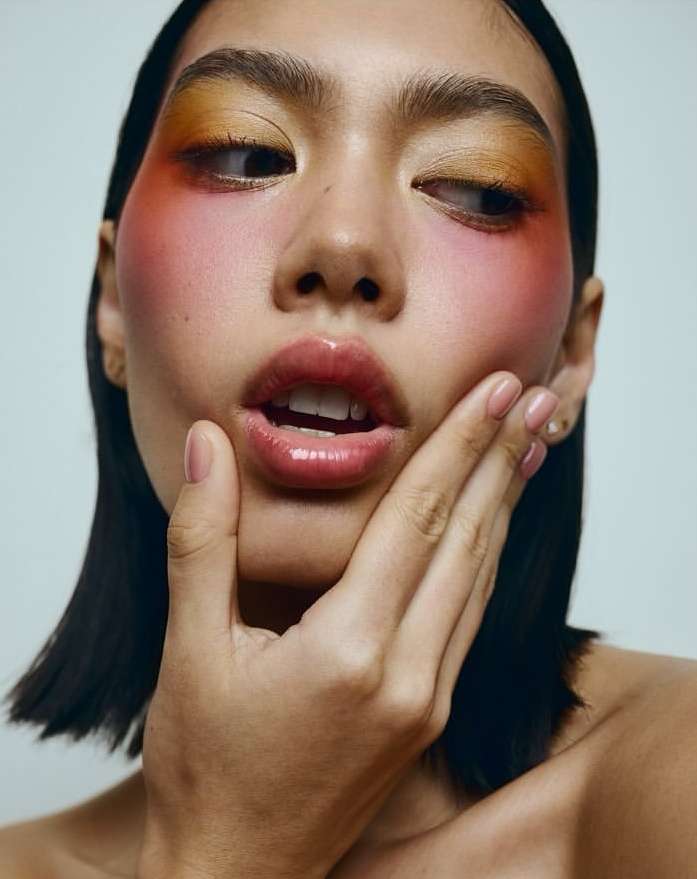 Close-up portrait of a woman with vibrant yellow and pink eye makeup and glossy lips, holding her hand to her face.