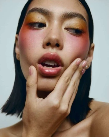 Close-up portrait of a woman with vibrant yellow and pink eye makeup and glossy lips, holding her hand to her face.